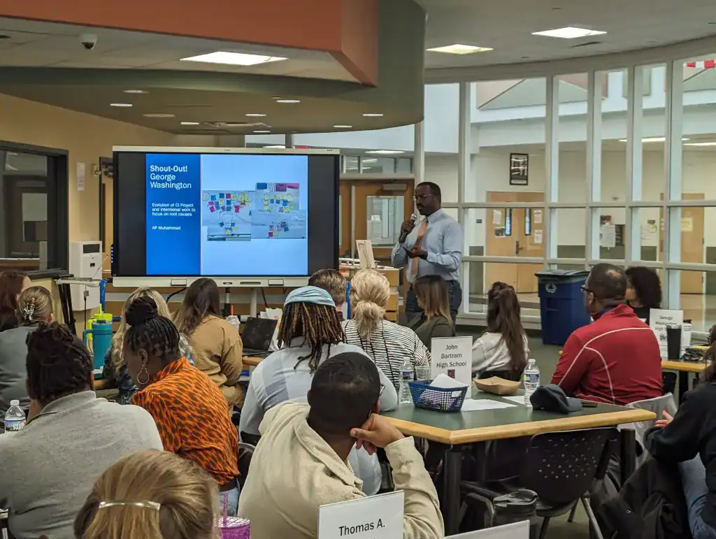 A man gives a lecture and other adults sit at tables to listen as part of the 9th Grade Success Network.