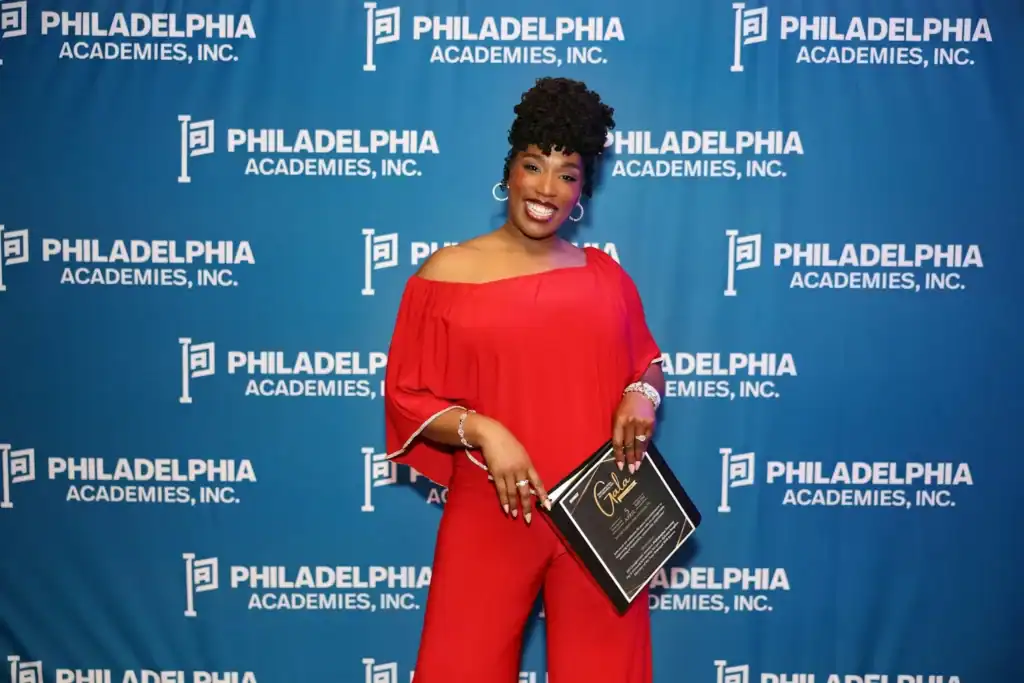 Woman standing in front of a Philadelphia Academies step and repeat holding a binder that contains information about their annual gala.
