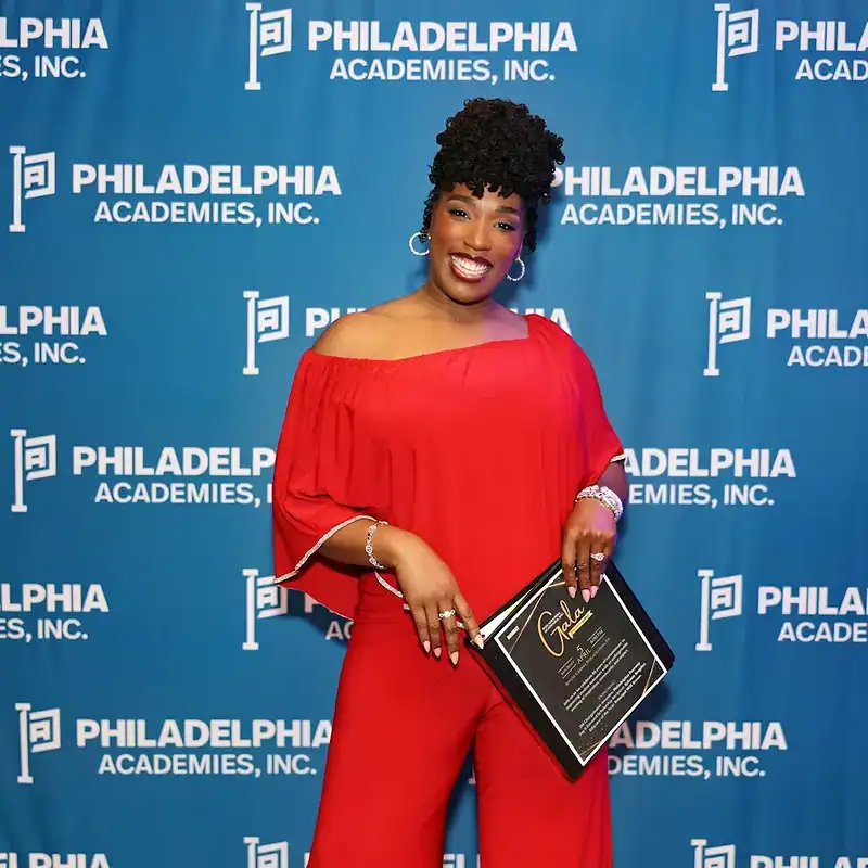Woman standing in front of a Philadelphia Academies step and repeat holding a binder that contains information about their annual gala.