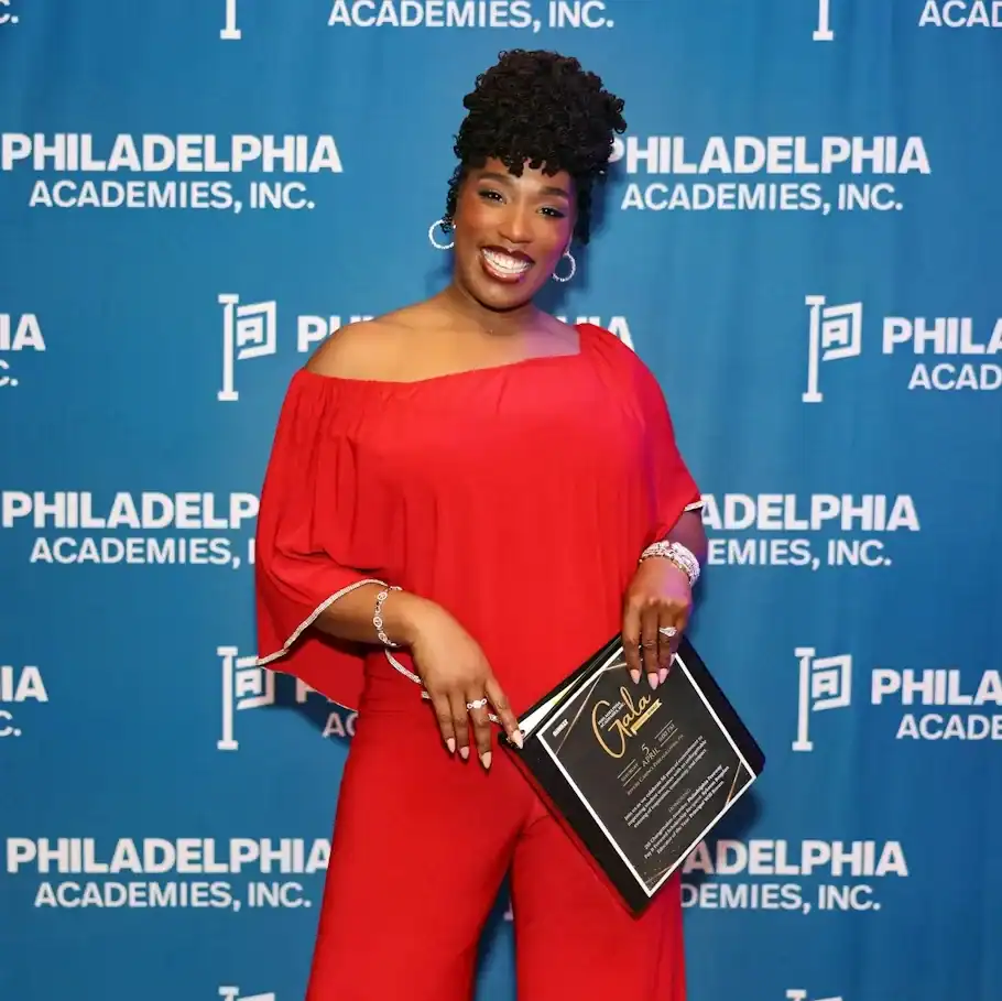 Woman standing in front of a Philadelphia Academies step and repeat holding a binder that contains information about their annual gala.