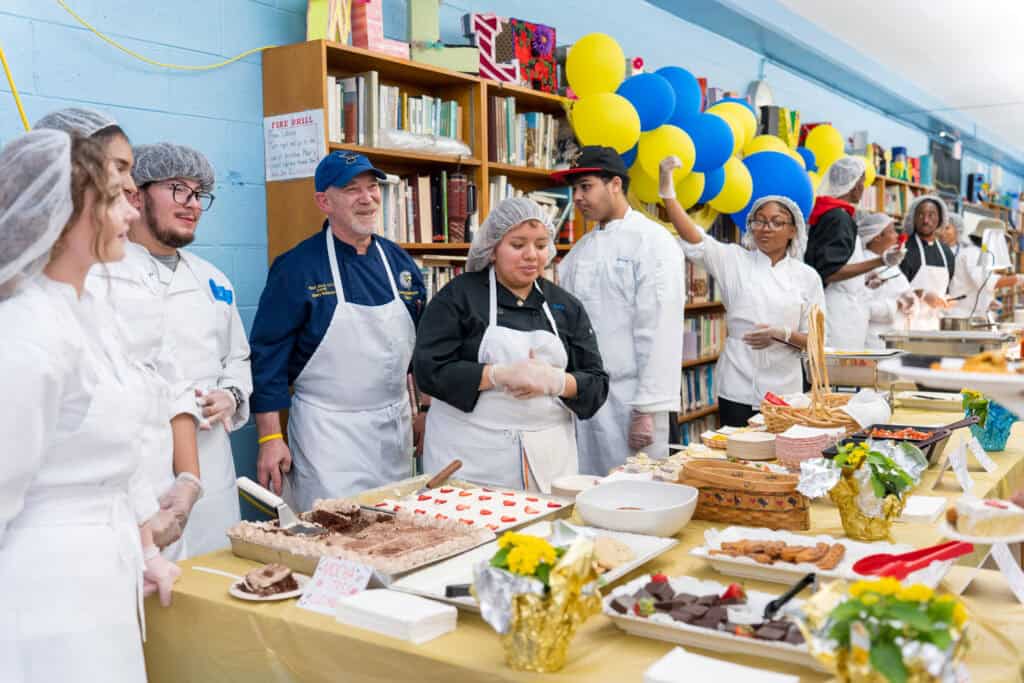 Staff in aprons and hairnets gather around tables of food to serve to the alumni network. There are yellow and blue balloons in the background. 