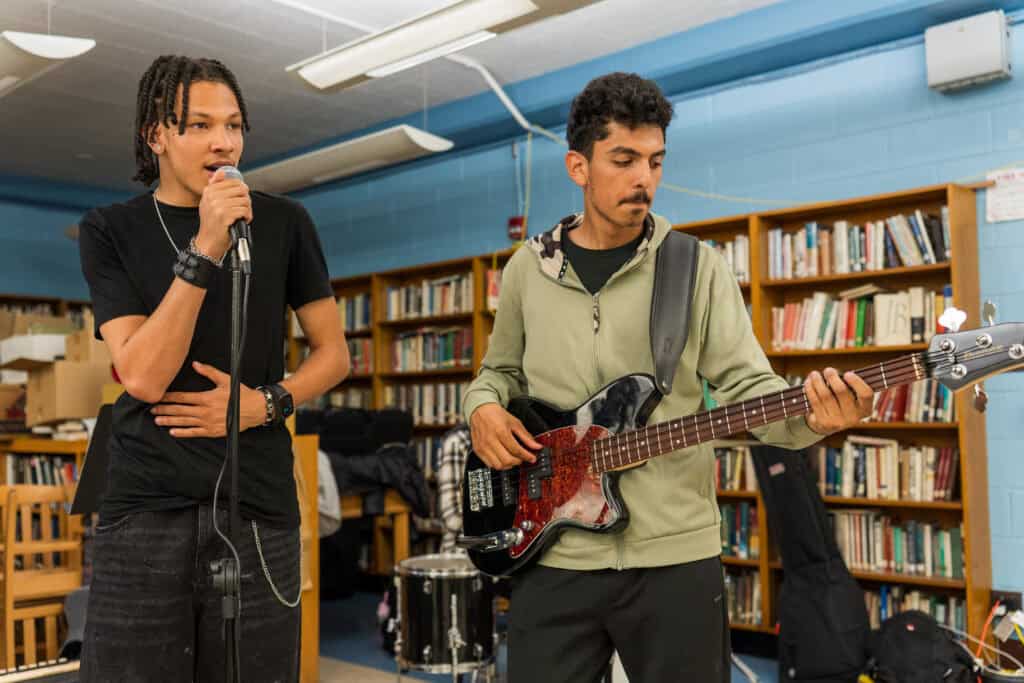 Two students perform in a band. One is singing and the other is playing electric guitar. They are inside a library.