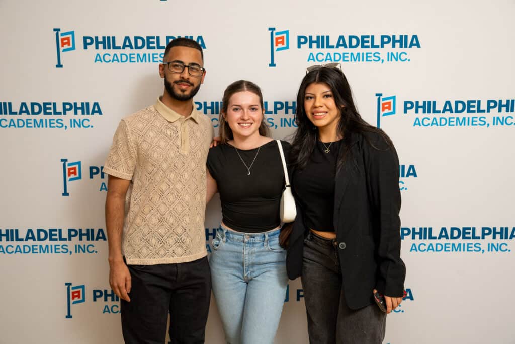 A man on the left, a woman in the middle, and a woman on the right stand in front of a Philadelphia Academies step and repeat. These students are a part of the Alumni Network.
