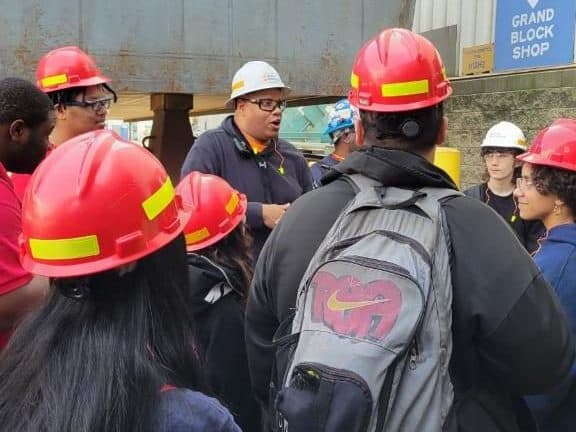 A man wearing a hardhat and protective glasses is guiding students wearing hardhats as well. This is a part of the workforce development programs.