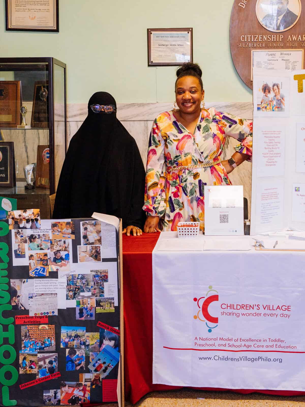 Two women stand beside one another. They are behind booths exhibiting information about Children's Village and early childhood education.