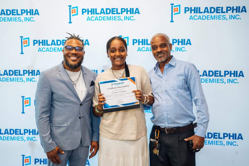 Two men stand beside a girl who is holding up a certificate in front of a Philadelphia Academies step and repeat. All parties are smiling.
