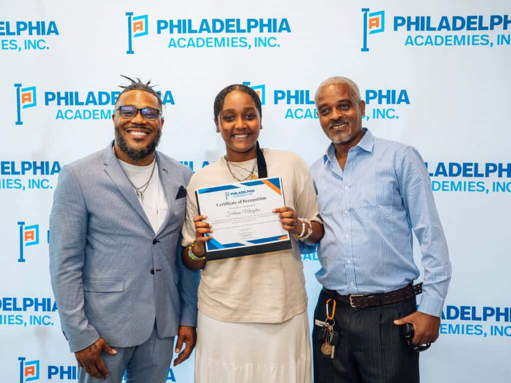 Two men stand beside a girl who is holding up a certificate in front of a Philadelphia Academies step and repeat. All parties are smiling.

