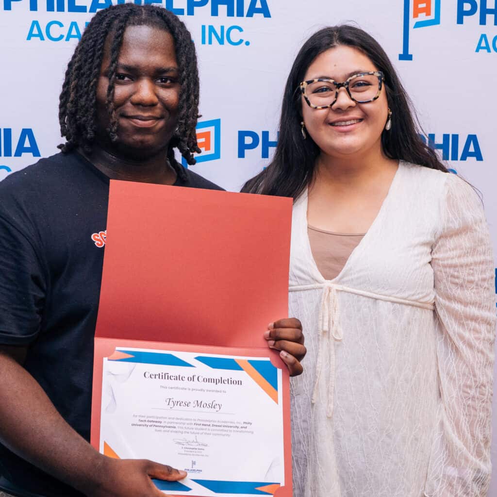 Student shows his certificate of completion in front of a Philadelphia Academies step and repeat. He's standing next to a smiling woman.