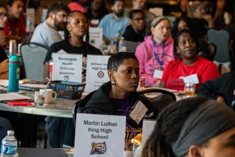 Representatives from different schools sit at tables and look to a screen as part of the 9th Grade Success Network.